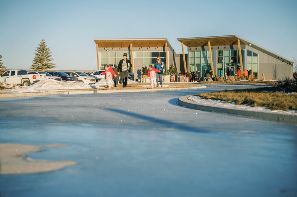 People walking and skating on an outdoor ice path in winter near a modern building with large glass windows and wooden beams.