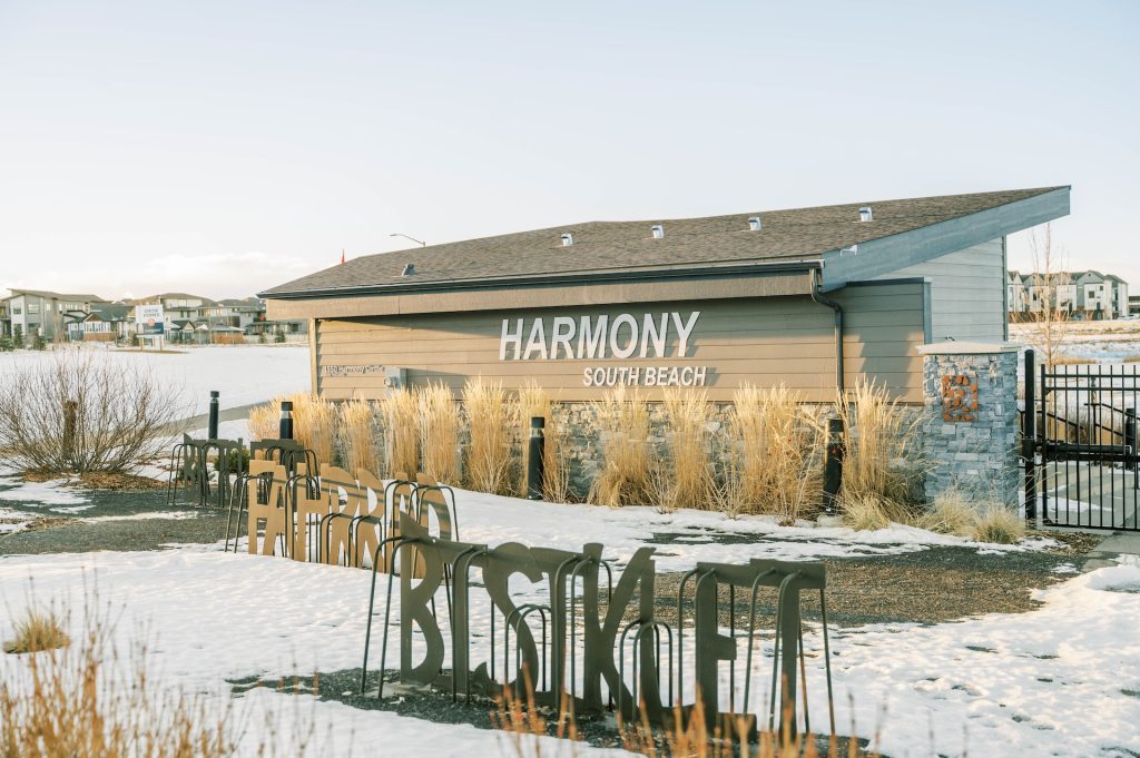 Harmony South Beach building with large signage, stone pillars, and ornamental grasses, surrounded by snow and nearby homes.